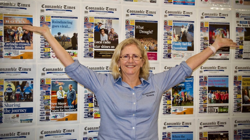 A woman stands with arms spread out in front of a wall of newspaper covers
