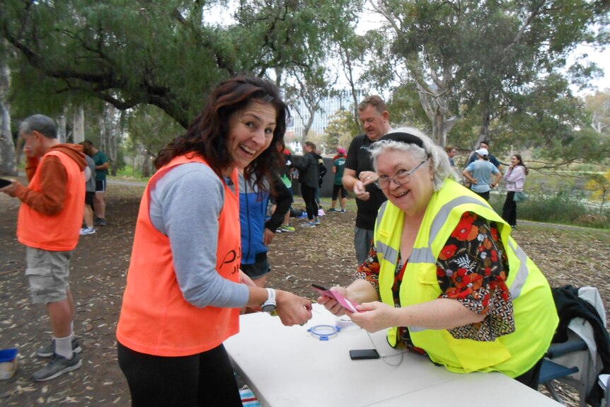 Merrilyn McMillan, dressed in a yellow high-vis vest, smiles while greeting a parkrun participant