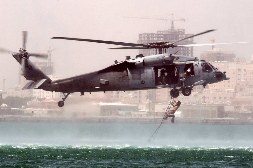 An image of a US grey helicopter hovering above a green sea with a man climbing a ladder touching the waterline underneath it.