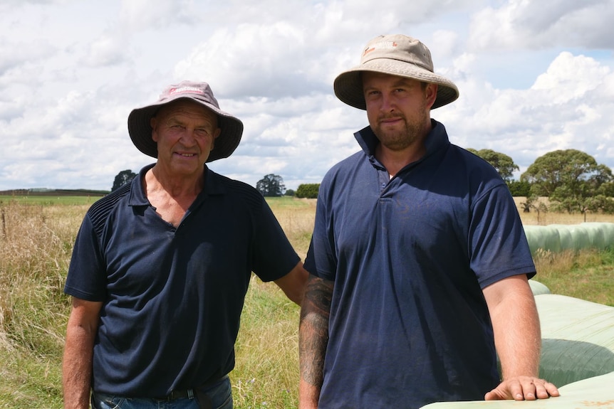 Two men in blue shirts and hats standing next to hay bales.
