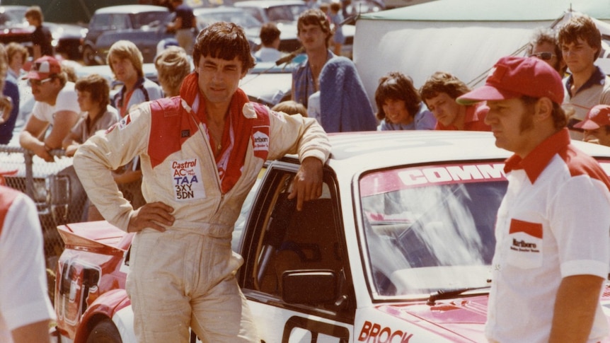 A man in overalls leans on a car with Brock written on it.