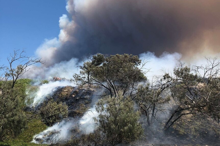 A large plume of smoke coming from behind a small hill covered in burnt scrub.
