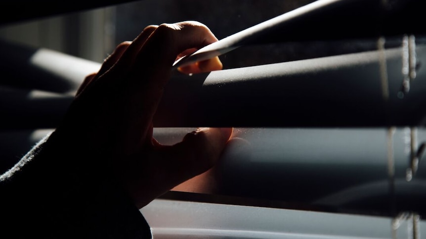 A hand is shown parting venetian blinds to enable a person to look out a window.