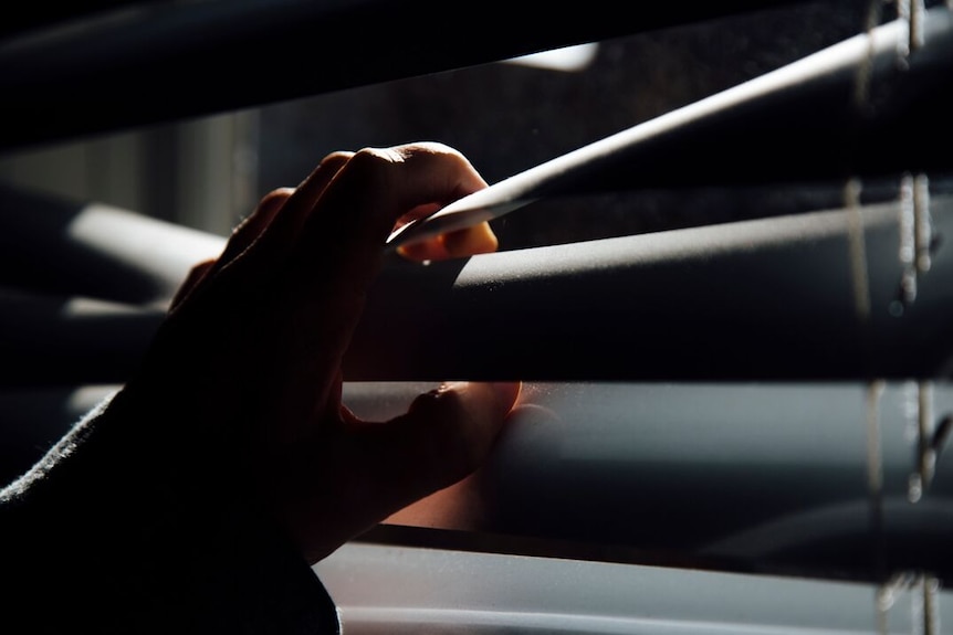 A hand is shown parting venetian blinds to enable a person to look out a window.