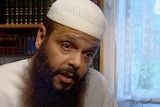 Man wearing white Islamic cap with long brown beard sits in study with books behind and window with laced curtains behind