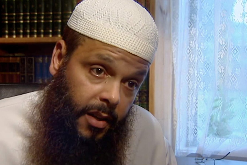 Man wearing white Islamic cap with long brown beard sits in study with books behind and window with laced curtains behind