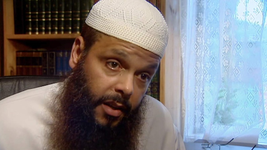 Man wearing white Islamic cap with long brown beard sits in study with books behind and window with laced curtains behind