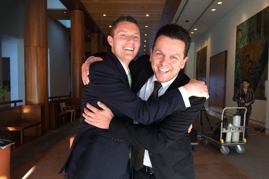 Two men in suits smile and embrace while looking at the camera