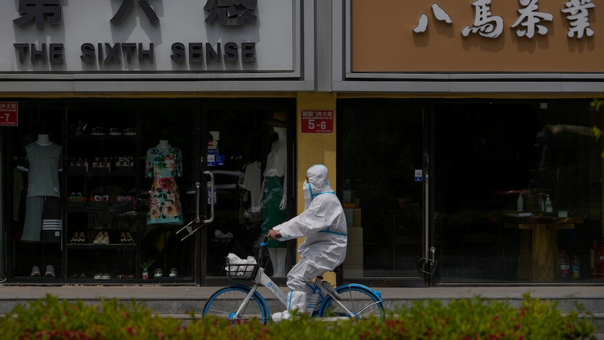 A worker in a protective suit rides a bicycle past shuttered shops