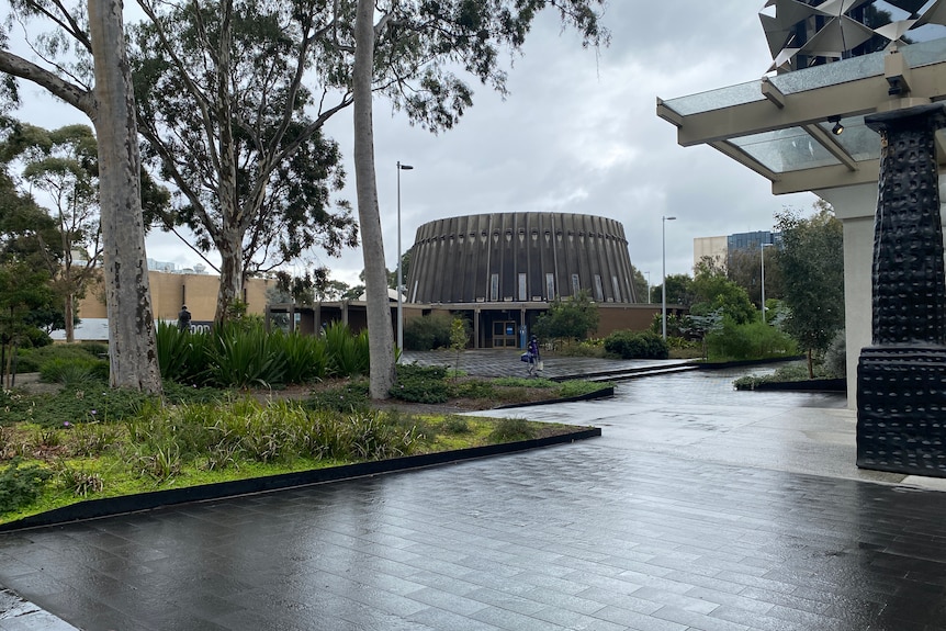 Building at a university with a green garden bed, wet road and rounded building in the background.