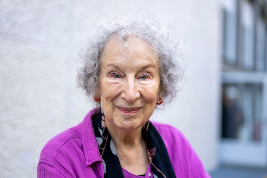 An elderly white woman with curly silver hair wearing a purple cardigan looks at the camera.
