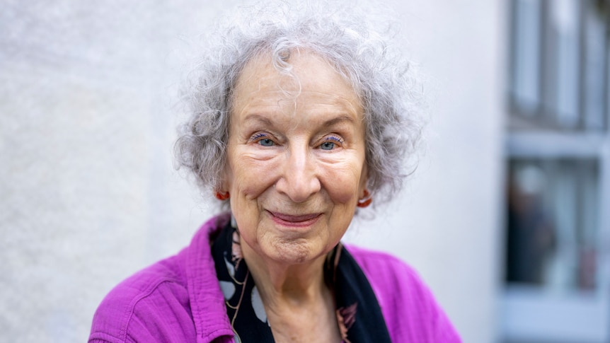 An elderly white woman with curly silver hair wearing a purple cardigan looks at the camera. 