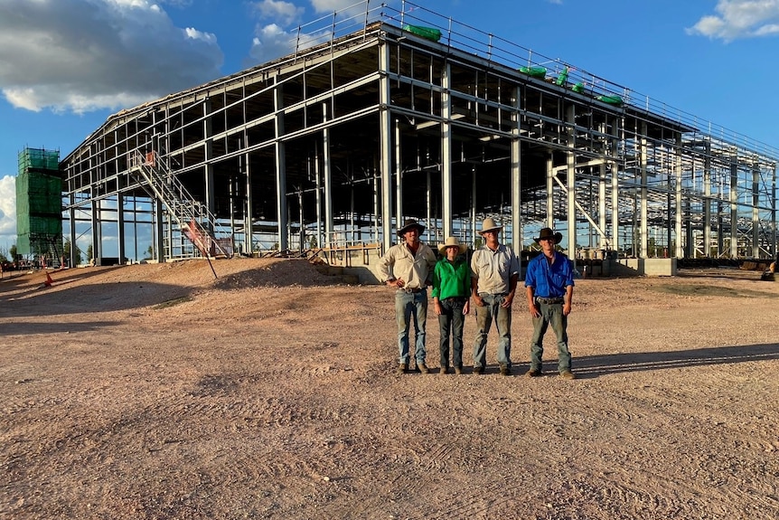 Four people stand on dirt, wearing hats, in front of large structure under construction, blue sky.