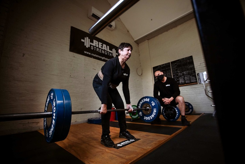 A woman lifts 100kg barbells in a gym and a man wearing a facemask watches