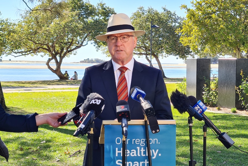 Man in suit with a hat on stands under trees at a podium with microphones