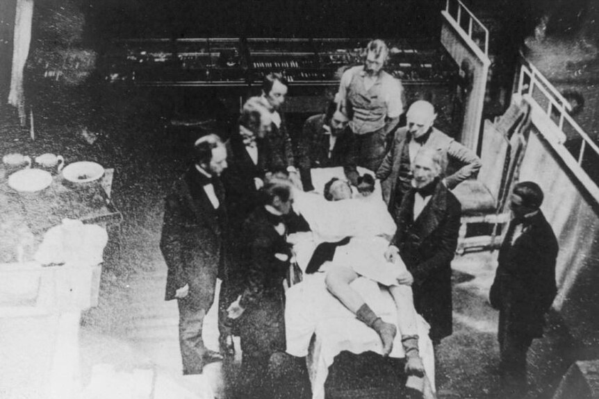 Old black and white photo of suited men standing around a patient on a hospital bed with sheet laying across him and bare legs.