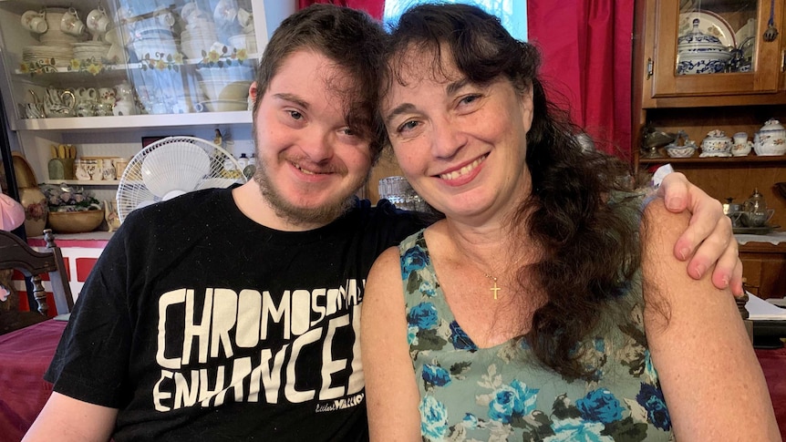 A man and his mum sit side-by-side, leaning their heads together as they smile at the camera