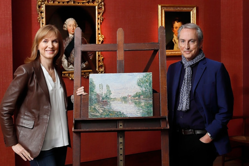 A man and a woman post next to a paintint on an easel in a red-walled gallery.