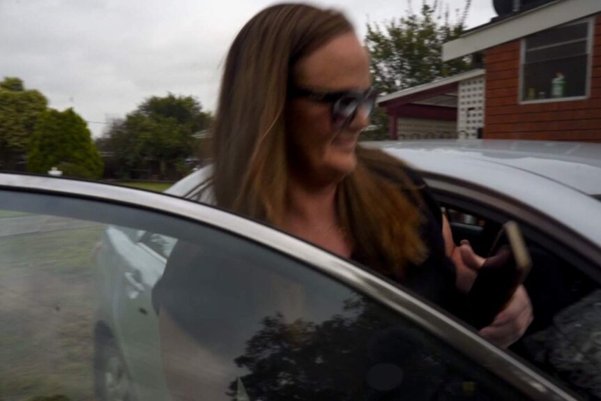 Woman with brown hair wearing sun glasses steps inside silver car with brick home and trees in background