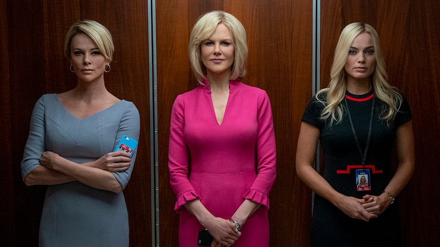 Three blonde women in office attire stand inside a walnut coloured lift, two look serious while one has slight smile.
