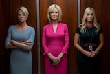 Three blonde women in office attire stand inside a walnut coloured lift, two look serious while one has slight smile.