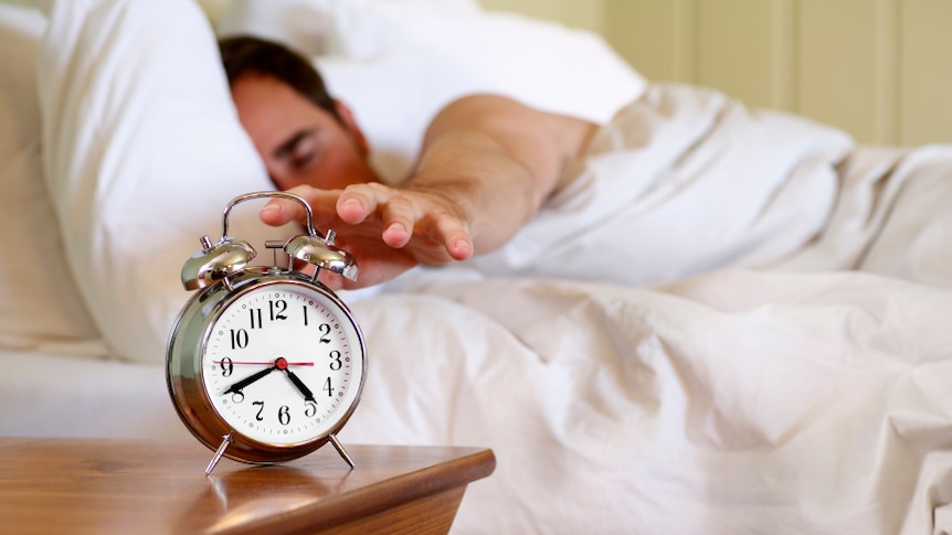 A man reaching out for an alarm clock while still in bed.