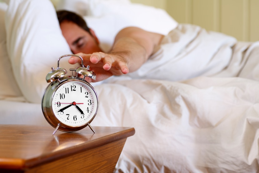 A man reaching out for an alarm clock while still in bed.