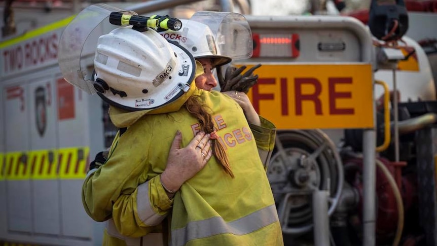 Two firefighters hug in front of a fire truck.