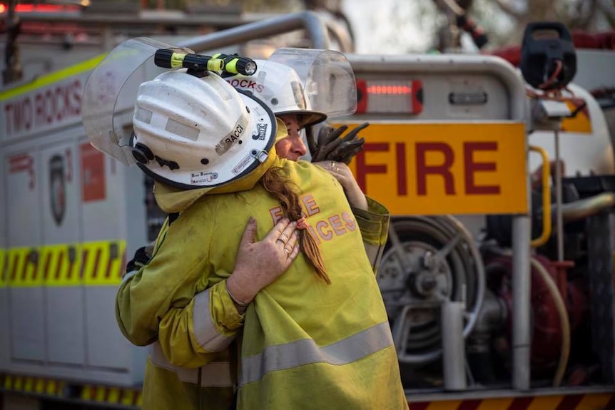 Two firefighters hug in front of a fire truck.