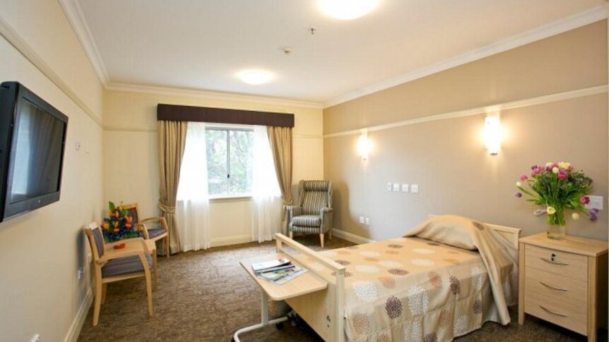 A room at an aged care facility with a bed, three chairs, a television and a bedside table with flowers