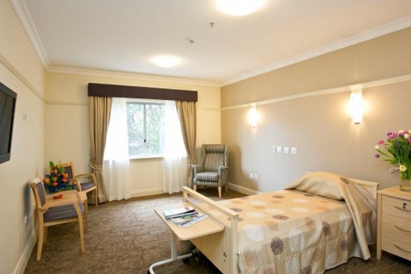 A room at an aged care facility with a bed, three chairs, a television and a bedside table with flowers.