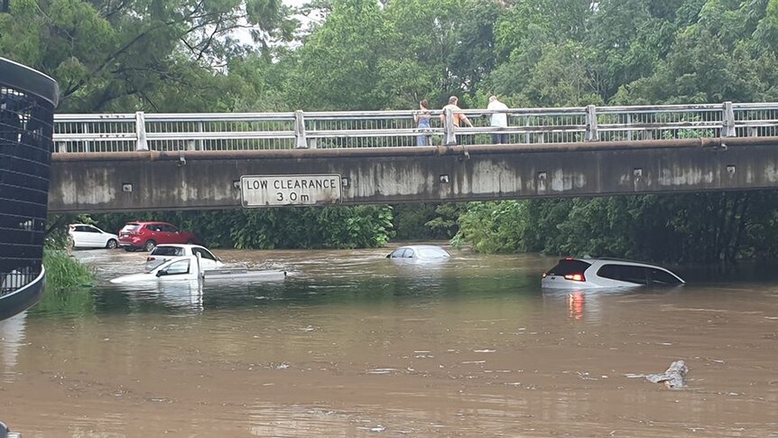 People look from an overpass at several cars submerged in floodwaters.