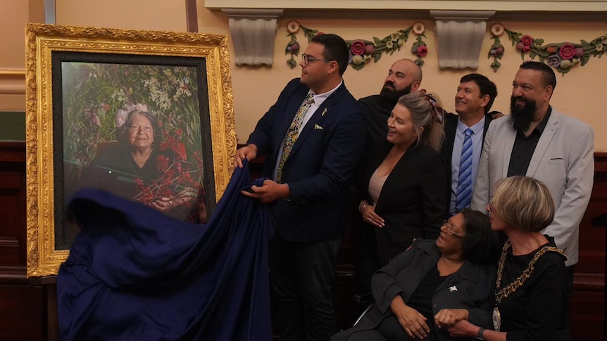 A woman in a wheelchair surrounded by family members looking at a large portrait of her with a thick golden frame