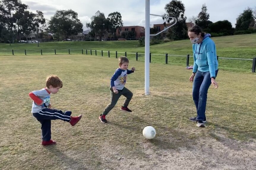 A woman wearing a face mask plays soccer with two young boys.