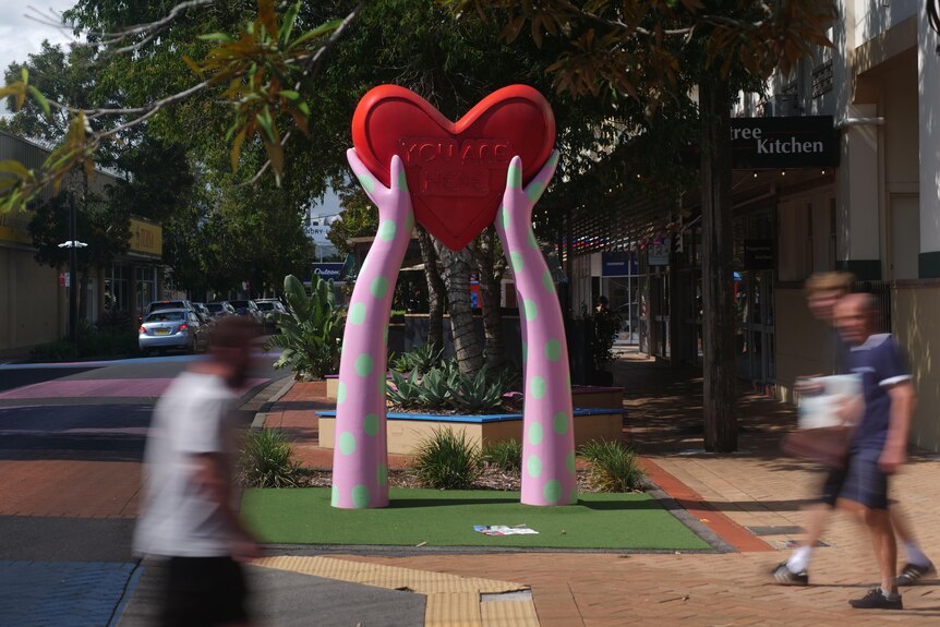 A statue of stylised hands holding a heart labelled you are here, with people walking past.