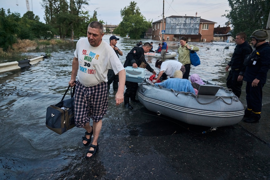 A man carries a bag as others behind him get into inflatable rafts as waters rise. 