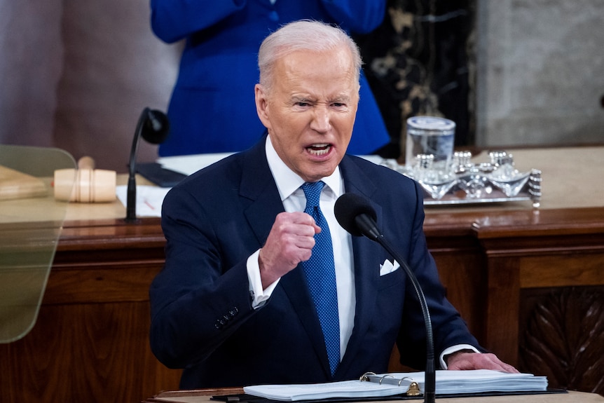 Joe Biden raises his fist while speaking at a lectern, looking angry 