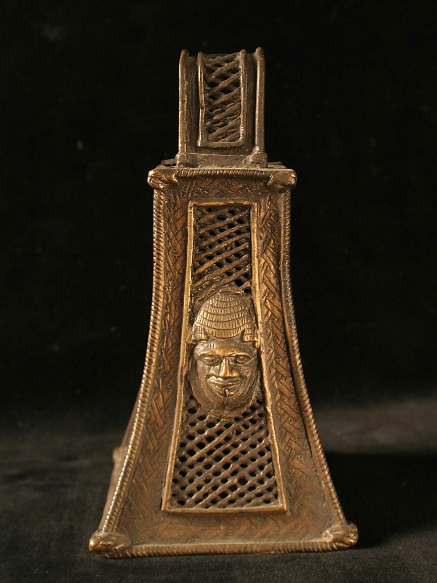 A four-sided brass bell decorated with a lattice pattern and on one side, a human face.