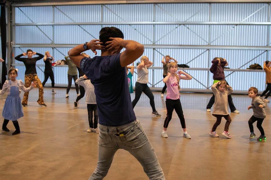 A man with his back to the camera instructs two rows of children and adults in Bollywood dance moves