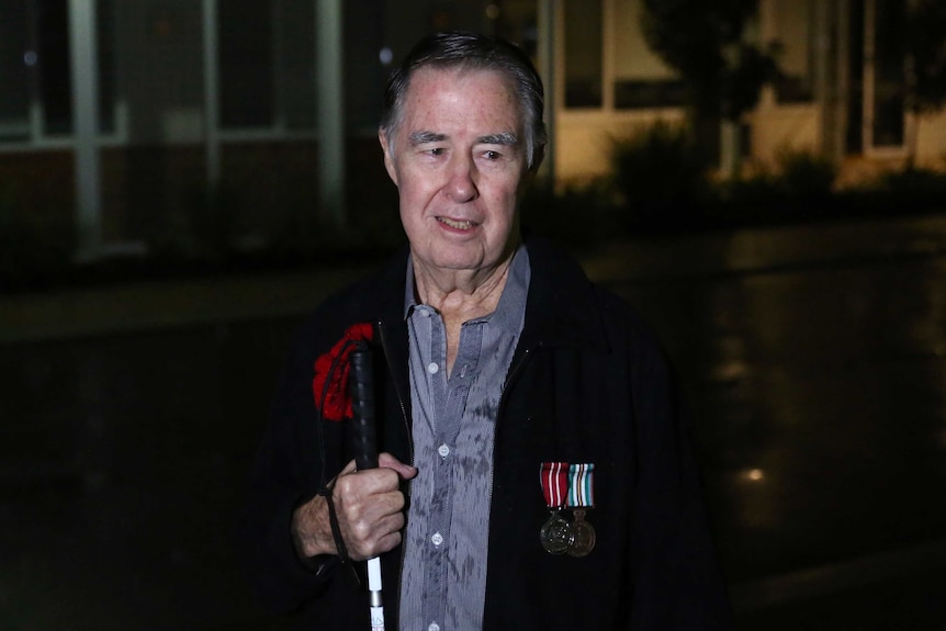 A man with a walking cane and service medals stands on a driveway