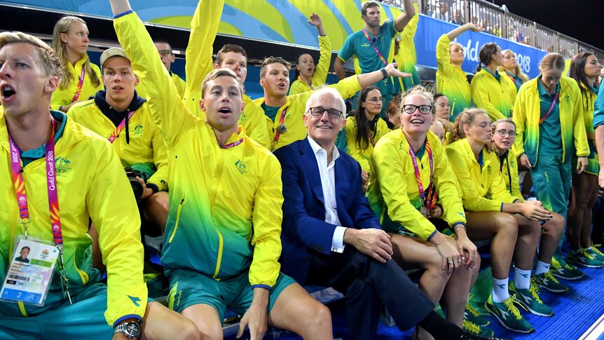 Malcolm Turnbull, wearing a suit, sits with hands folded on his lap, among swimmers in Commonwealth Games tracksuits cheering.