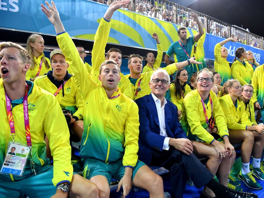 Malcolm Turnbull, wearing a suit, sits with hands folded on his lap, among swimmers in Commonwealth Games tracksuits cheering.
