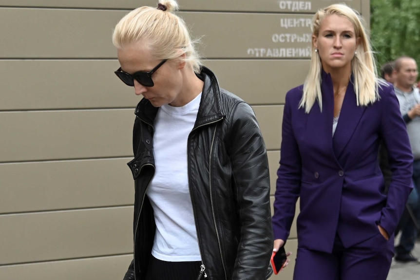 A woman in a white top and black leather jacket and dark glasses walks ahead of another woman in a suit.