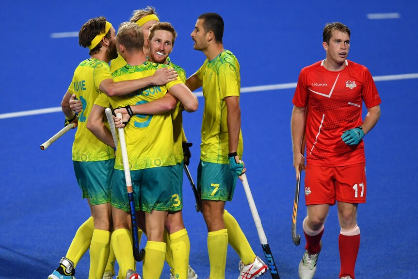 A group of Australian men's hockey players hug after a goal, as a dejected England player stands by.