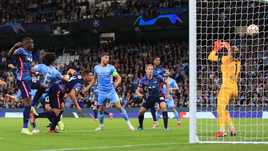 A Manchester City attacker falls between two defenders as the ball flies into the net from his header.