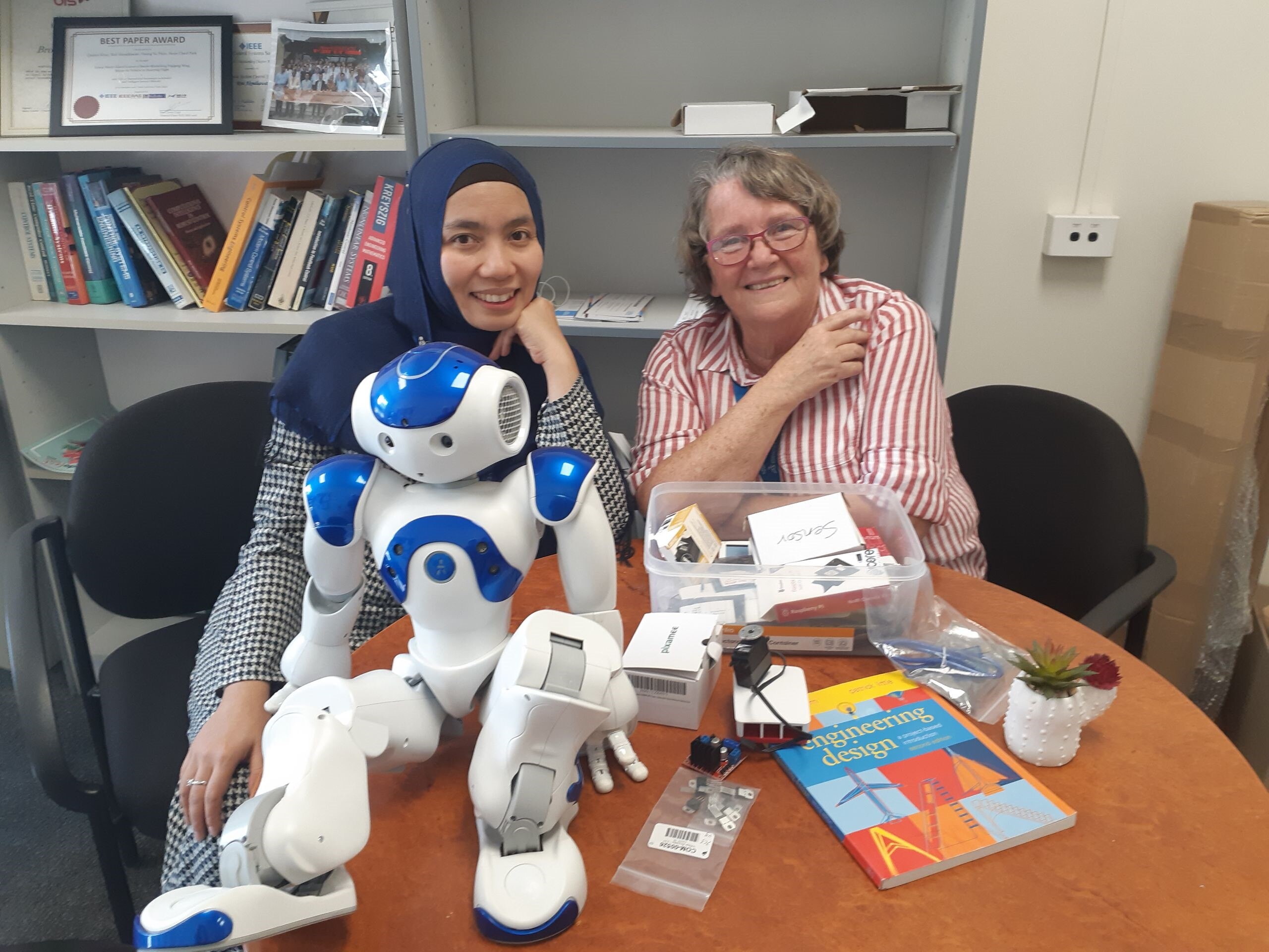 Rini sitting down in front of a robotic model dan books and her colleague