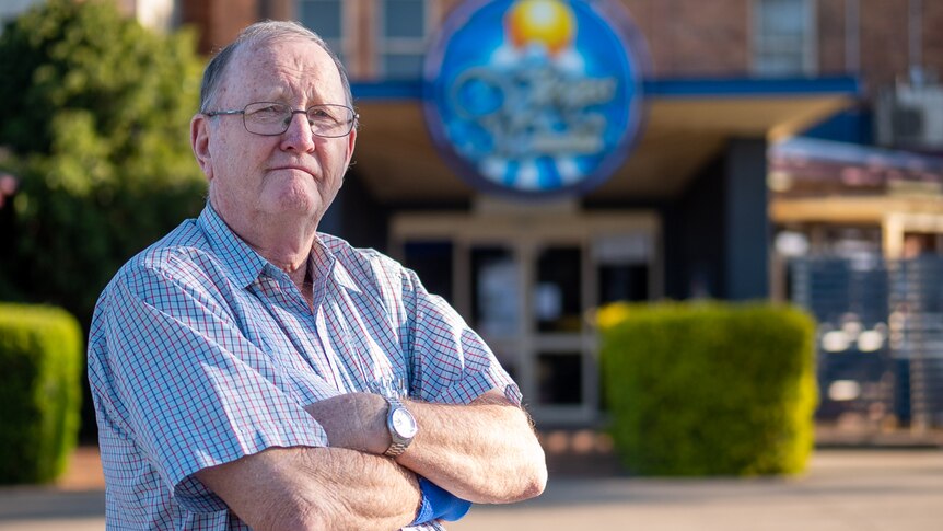 Older man wearing glasses stands arms folded outside with building and green hedge visible in background