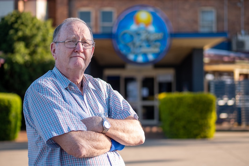 Older man wearing glasses stands arms folded outside with building and green hedge visible in background