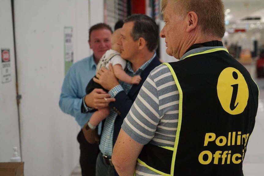 A man holding a baby stands near another man with a vest that reads 'polling officer' on the back