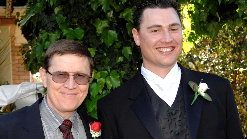 Two men, one in glasses, wearing suits and flower lapels, at a wedding.
